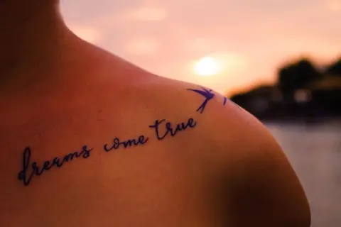 Schulter mit Tattoo "dreams come true" in geschwungenen Tattoo Schriftarten, daneben eine kleine Schwalbe.