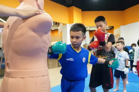 A young boy in a blue shirt practices a boxing feint, throwing a punch at a training dummy while other children watch.