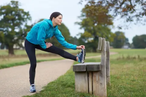 Donna in abbigliamento sportivo blu esegue un riscaldamento pre corsa, allungando la gamba su una panchina in un parco.
