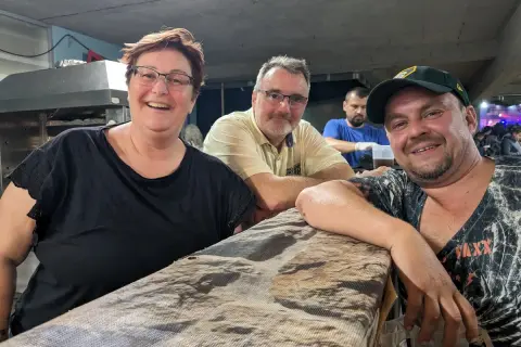 Fröhliche Menschen am holzmarkt ilsfeld. Eine Frau mit Brille und schwarzem Shirt, ein Mann mit gelbem Hemd und ein Mann mit Kappe lächeln in die Kamera.