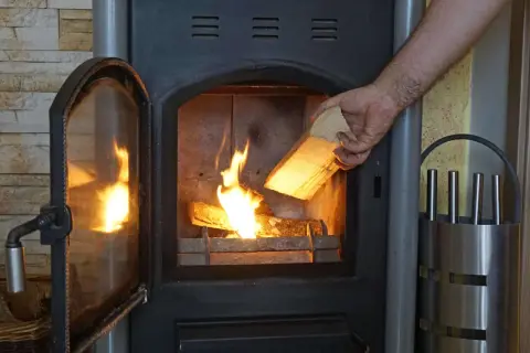 Hand legt Holzscheit mit gutem Brennwert Birke in den brennenden Ofen.
