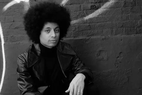 Jose James z burzą afro, w skórzanej kurtce i golfie, siedzi na tle ściany z graffiti.