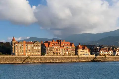 Edificios históricos con tejados rojos bordean el paseo de las grandes villas Getxo, con montañas al fondo y el mar en primer plano.