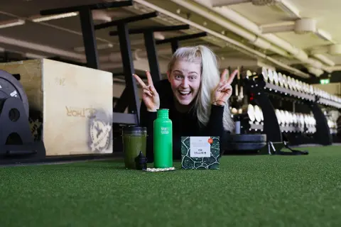Glad kvinna med energi i gymmet, redo att ta sig an dagen. Frågan är: hur blir man pigg? Kanske med en grön smoothie och kosttillskott?