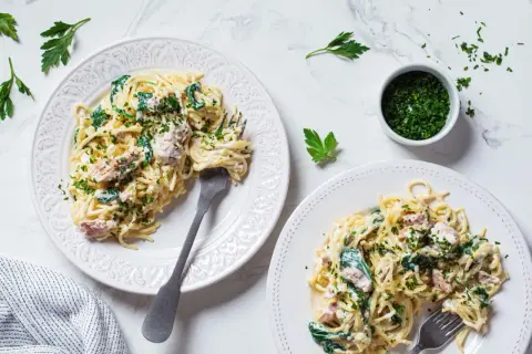 Dwa talerze spaghetti z kremowym sosem, szpinakiem i kawałkami tuńczyka z puszki, posypane natką pietruszki.