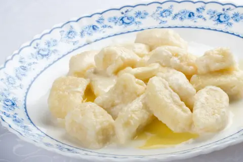 Pyszne kopytka z serem, polane śmietaną i masłem, na talerzu z niebieskim wzorem.