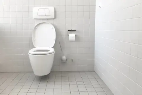 Bagno bianco con WC aperto. Si nota una piccola macchia d'acqua sul pavimento, forse il galleggiante wc perde acqua.