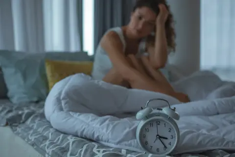 Kobieta siedzi na łóżku, trzymając się za głowę. Budzik obok pokazuje wczesną godzinę. To klasyczne objawy niewyspania.