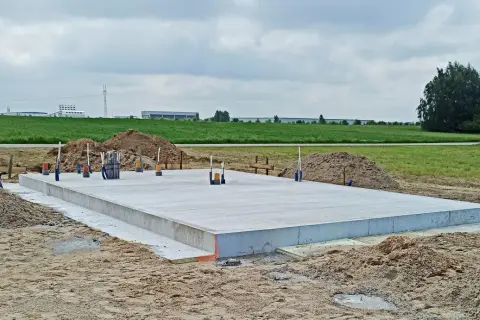 Gotowa płyta fundamentowa na budowie, otoczona piaskiem i zielenią. W tle widać budynki przemysłowe.