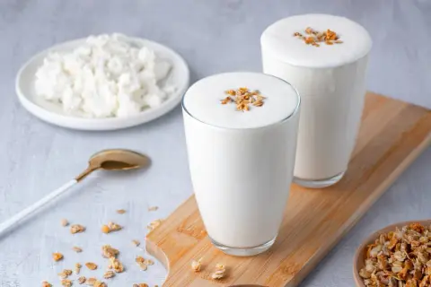Deux verres de kefir de lait crémeux, garnis de granola, à côté d'une assiette de fromage blanc et d'une cuillère dorée.