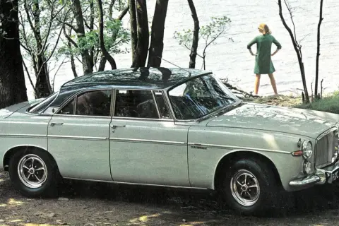 En ljusgrå Rover P5 står parkerad vid en sjö. En kvinna i grön klänning står vid vattenbrynet.