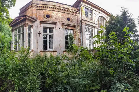 Zarośnięty dworek zakoniczyn z ceglanymi fragmentami i dużymi oknami, otoczony bujną zielenią.