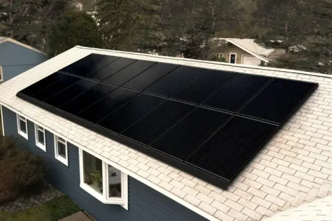 Paneles negros de una aplicacion placas solares cubren el tejado de una casa azul, rodeada de árboles.