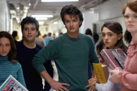 A group of teens in a hallway, featuring the iconic Stranger Things color palette of muted blues, greens, and pinks.