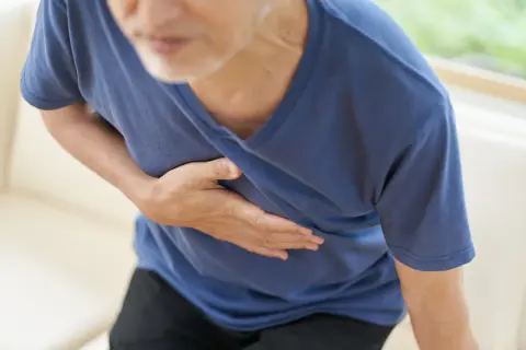 Un homme âgé se tient la poitrine, le visage marqué par la douleur. Sa main droite serre son cœur, comme s'il ressentait une douleur vive, une côte félée peut-être.