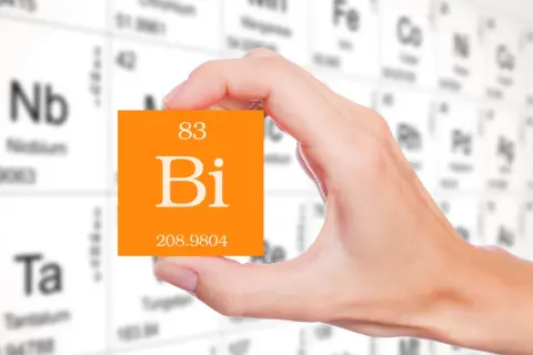 Dłoń trzyma pomarańczową kartkę z symbolem pierwiastka bizmut (Bi) i jego liczbą atomową 83. W tle widać tablicę Mendelejewa.