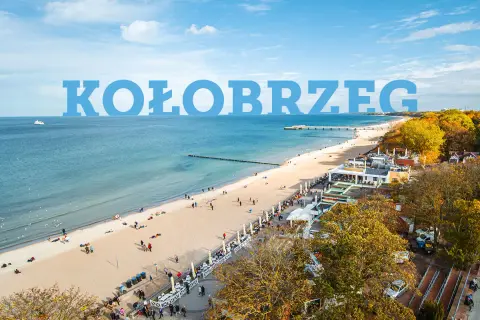 Widok na plażę w Kołobrzegu z ludźmi, budynkami i morzem.
