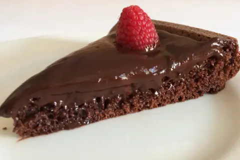 Una fetta di gateau al cioccolato, lucida e golosa, con una fragola fresca in cima, su un piatto bianco.