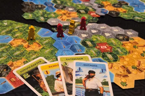 Spielbrett mit Figuren und Karten. Die Karten "KUNDSCHAFTER", "REISEN", "ENTDECKER" und "MATROSE" sind ausgelegt. Eine deutsche Spielepreis Liste könnte diese Elemente zeigen.