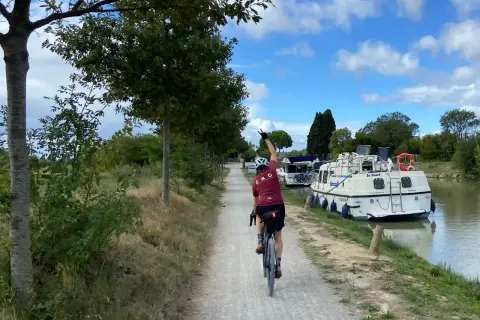 Ciclista pedaleando por el Canal du Midi, con barcos amarrados a un lado y árboles al otro.