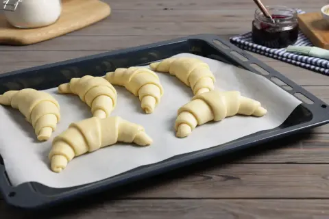 Sześć surowych rogalików z ciasta francuskiego czeka na pieczenie. Obok słoiczek z dżemem.