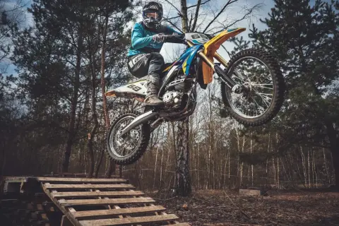 Motocyklista w kasku i goglach skacze na motocyklu enduro nad drewnianą rampą w lesie.