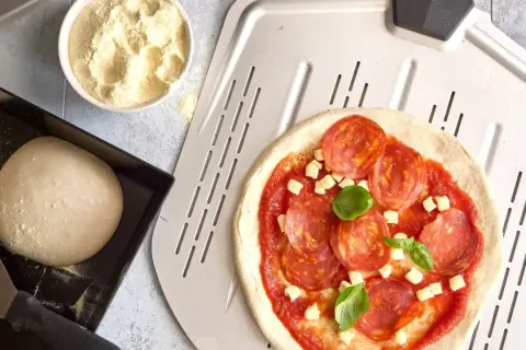 Pizzadeg recept: färdig pizzadeg, tomatsås, salami och ost på en pizzaspade. Bredvid en skål med mjöl och en form med pizzadeg.