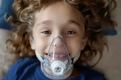 Uśmiechnięte dziecko z kręconymi włosami w masce inhalacyjnej.