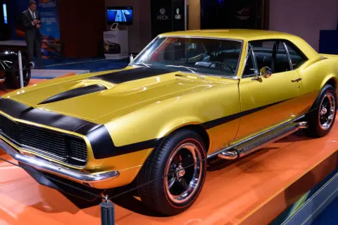 En glänsande gul Chevrolet Camaro SS 1969 med svarta ränder på huven och sidan. Bilen står på en orange yta.