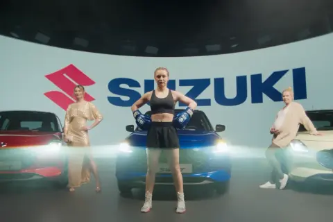 Trzy kobiety obok samochodów Suzuki. Jedna w złotej sukni, druga w stroju bokserskim, trzecia w ciąży. Swift język programowania inspiruje do dynamicznych zmian.