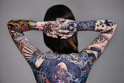 Rückenansicht einer Frau mit aufwendigen, abstrakten Tattoos, die ihren ganzen Körper bedecken. Goldene Nägel und Schmuck ergänzen die Kunstwerke.