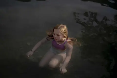 Bambina con capelli biondi immersa nell'acqua scura, un'immagine che evoca la serenità e la purezza, tipica delle opere di Laura Pannack.