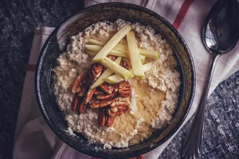 Porridge zubereiten: Cremiger Haferbrei mit Apfelspalten, Pekannüssen und Honig in einer Schüssel, daneben ein Löffel.
