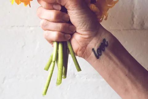 Ein Handgelenk mit einem "Love"-Tattoo, das an ein Freundschaftstattoo erinnert, hält einen Strauß gelber Blumen.