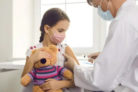 Dziewczynka z maską na twarzy i pluszowym misiem przytula się do ramienia, podczas gdy lekarz wykonuje szczepienia covid.