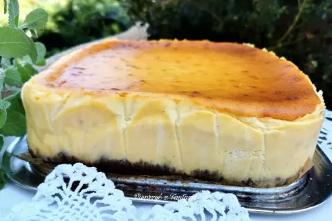 Pyszny sernik z mleka w proszku, z lekko przypieczoną skórką, na metalowej tacy, otoczony zielenią i białą koronką.