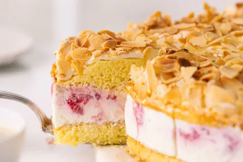 Ein Stück himmelstorte mit himbeeren wird mit einer Gabel herausgehoben. Die Torte ist mit Mandelsplittern bestreut.