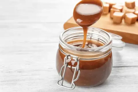 Une cuillère verse un coulis de caramel onctueux dans un bocal en verre. Des bonbons au caramel sont disposés sur une planche en bois en arrière-plan.