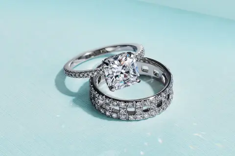 Un anillo de compromiso con un gran diamante cuadrado reposa sobre un anillo de matrimonio con incrustaciones de diamantes, ambos de plata.
