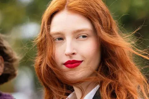 Eine Frau mit leuchtend roten Haaren, die im Wind wehen. Ihr Blick ist nach rechts gerichtet, ihre Lippen sind rot geschminkt. Natur und rote Haare vereinen sich in diesem Porträt.