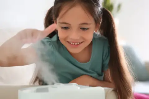 Uśmiechnięta dziewczynka bawi się mgiełką z nawilżacza powietrza dla niemowląt, tworząc magiczną atmosferę w pokoju.
