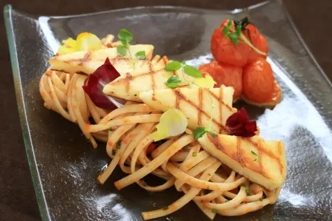 Makaron z halloumi, grillowane plastry sera, pomidorki koktajlowe i listki ziół na szklanym talerzu.