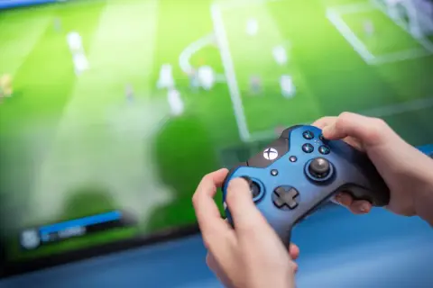 Ręce trzymające niebieski kontroler Xbox, grające w grę piłkarską. Ekran telewizora pokazuje zielone boisko z zawodnikami, tworząc efektowną teselację.