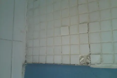 Carrelage blanc qui se décolle, craquelé et endommagé par l'humidité. Des carreaux sont cassés, révélant le mur derrière.