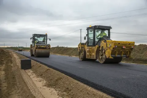 Dwa żółte walce drogowe, każdy to wolnobieżny pojazd budowlany, ubijają świeży asfalt na nowej drodze.