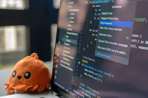 Un adorable crabe en peluche à côté d'un écran d'ordinateur affichant du code. Le menu contextuel propose des actions IA, aidant à choisir quel langage de programmation apprendre.
