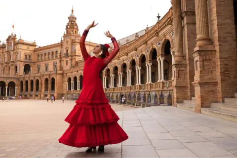 Bailarina con vestido rojo voluminoso, brazos en alto, ejecutando sevillanas lentas frente a un edificio histórico con arcos.