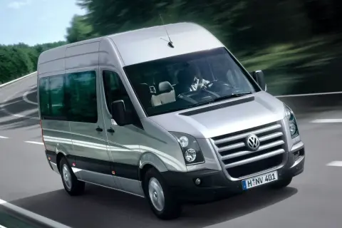 Srebrny Volkswagen Crafter jedzie po krętej drodze wśród drzew. Kierowca w białej koszuli prowadzi auto.