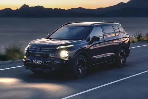 Ciemny Mitsubishi Outlander PHEV jedzie drogą o zachodzie słońca, z górami i jeziorem w tle.