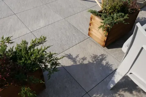 Nowe, szare płyty tarasowe ułożone na betonie, z drewnianymi donicami i zielenią.